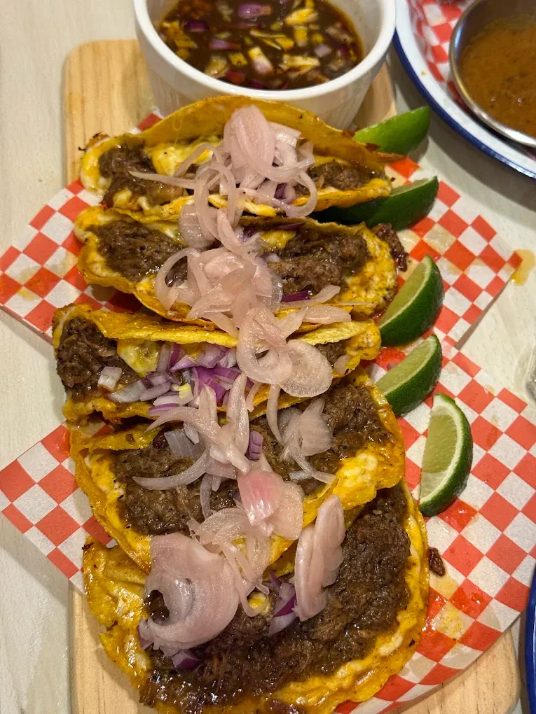 Five cheese-crisped birria beef tacos topped with pickled onions, with lime wedges and beef consommé