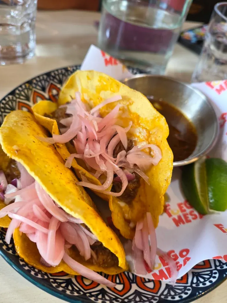 Two birria beef tacos in yellow corn tortillas topped with pickled onion, with beef consommé and lime alongside