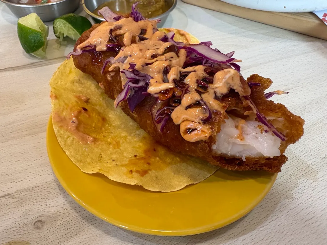 A single beer-battered fish taco on a yellow plate, showing flaky white fish under purple cabbage and chipotle mayonnaise
