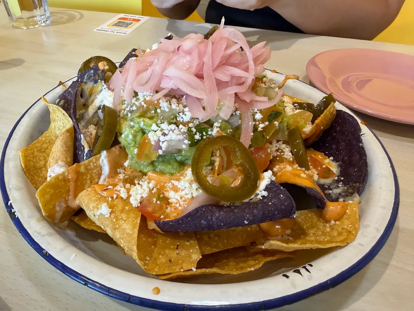 A bowl of loaded nachos built on blue and yellow corn chips with guacamole, jalapeños and pickled onions