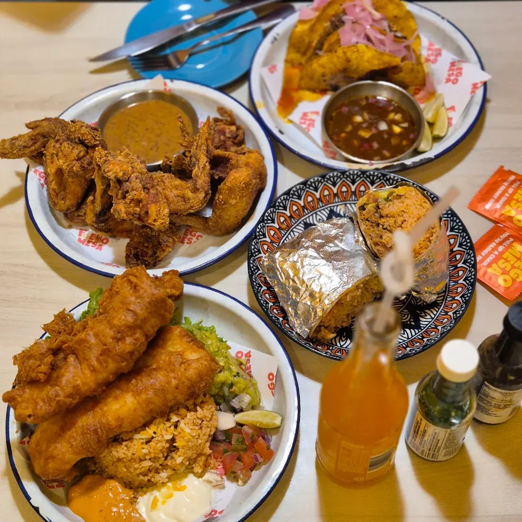 A shared table of fried chicken wings, birria tacos, a foil-wrapped burrito and battered fish with rice