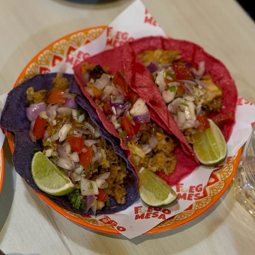 Three tacos on purple and pink corn tortillas with shredded filling, pico de gallo and lime wedges